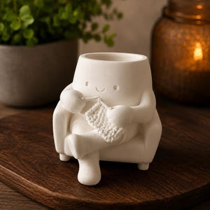May include: A matte white ceramic planter shaped like a character sitting in an armchair whilst knitting. The character has a simple smiling face and is holding a small piece of textured white knitting with a hook. The planter has a hollow top opening.