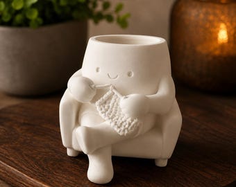 May include: A matte white ceramic planter shaped like a character sitting in an armchair whilst knitting. The character has a simple smiling face and is holding a small piece of textured white knitting with a hook. The planter has a hollow top opening.