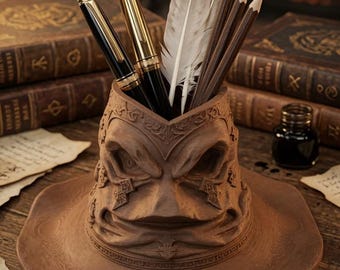 May include: A brown, sculpted desk organiser shaped like a weathered, wide-brimmed hat with deep facial features and intricate embossed patterns. It holds two fountain pens with gold accents, two grey-tipped feathers, and three dark wooden pencils.