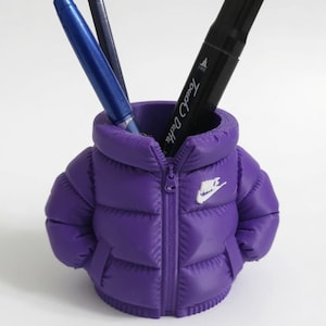 May include: A purple pencil holder shaped like a miniature puffer jacket with a white Nike logo on the chest. The jacket features a central zip detail, quilted texture, and two side pockets. Inside the holder are two pens and a pencil.