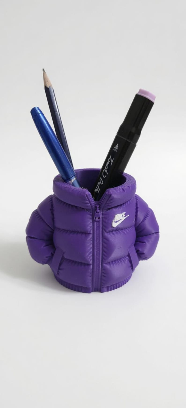 May include: A purple pencil holder shaped like a miniature puffer jacket with a white Nike logo on the chest. The jacket features a central zip detail, quilted texture, and two side pockets. Inside the holder are two pens and a pencil.
