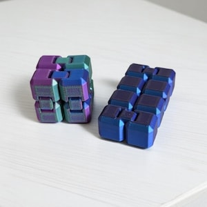May include: Two 3D-printed fidget puzzles with a metallic finish. One is a cube, the other is a rectangular shape. The colours shift from blue to purple to green. The word "Handmade" is in the top right corner. The text "Unique 3D Printed Puzzle" is at the bottom.