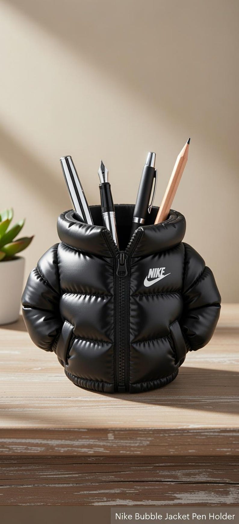 May include: A black puffer jacket shaped pen holder featuring a white Nike logo on the chest. The jacket has a central zip, a high collar, and two side pockets. Several pens and a wooden pencil are stored inside the open neck of the jacket.