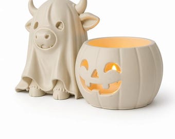 May include: A matte beige ceramic figurine of a cow wearing a ghost costume standing next to a matching matte beige ceramic pumpkin-shaped candle holder with a carved jack-o'-lantern face and a warm glowing light visible from the inside.