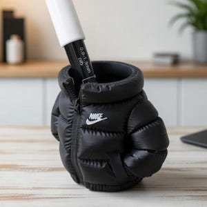 May include: A black puffer jacket shaped pen holder featuring a white Nike logo on the chest. The jacket has a zippered front design and contains a black marker with a white cap and text reading 0.9-1.3mm nib inserted into the top opening.
