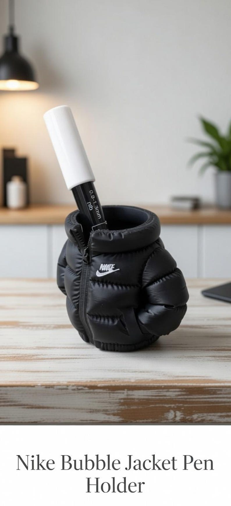 May include: A black puffer jacket shaped pen holder featuring a white Nike logo on the chest. The jacket has a zippered front design and contains a black marker with a white cap and text reading 0.9-1.3mm nib inserted into the top opening.