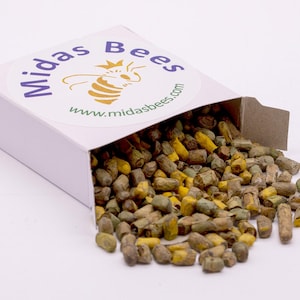 May include: A white box with the text "Midas Bees" and a bee logo, spilling out a pile of bee pollen. The pollen is a mix of brown, yellow, and green granules. The website address www.midasbees.com is also visible.