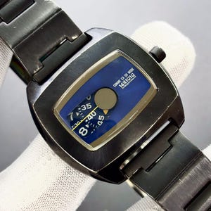 May include: A rectangular, dark gray wristwatch with a blue face. The watch face displays the time and the text "COMME CA DU MODE 1481010 Independent". The watch has a dark gray metal band.