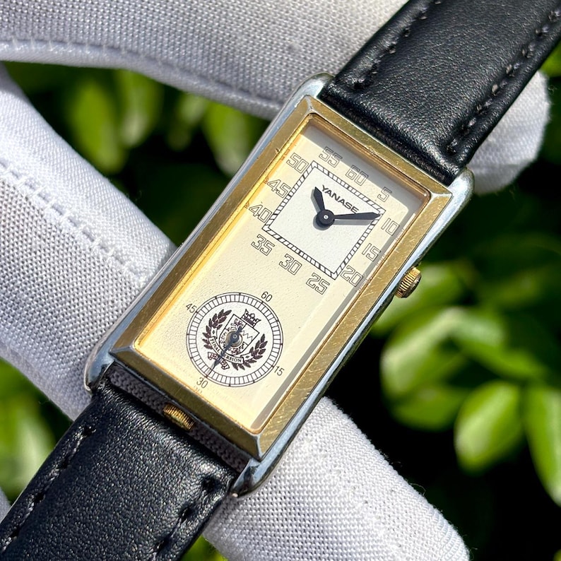 May include: A rectangular gold-toned wristwatch with a black leather band. The watch face is cream-colored with black numerals and hands. The brand name "YANASE" is visible. A circular emblem is at the bottom of the watch face.