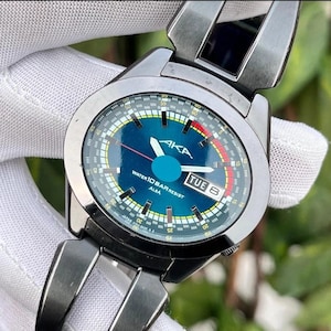 May include: A silver AKA Alba watch with a blue face and a matching strap. The watch face displays the day of the week, "TUE 8", and the text "WATER 10 BAR RESIST ALBA". The watch has a round face with white and red accents.