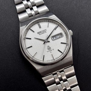 Pode incluir: Um relógio de pulso Seiko Quartz Type II prateado com mostrador branco texturizado, índices de bastão prateados e ponteiros prateados. O relógio possui uma janela de dia e data na posição das três horas e uma pulseira de aço escovado.
