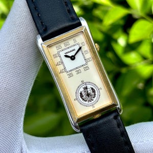 May include: A rectangular wristwatch with a gold-tone bezel and a black leather band. The watch face is cream-colored with black numerals and hands. The brand name "YANASE" is visible on the face, along with a crest emblem.