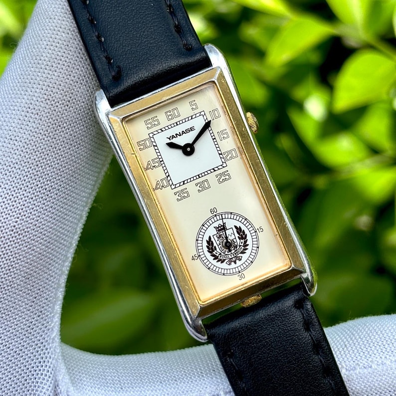 May include: A rectangular wristwatch with a gold-tone bezel and a black leather band. The watch face is cream-colored with black numerals and hands. The brand name "YANASE" is visible on the face, along with a crest emblem.