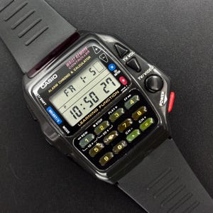 Op de afbeelding: Zwarte Casio digitale horloge met rekenmachine- en afstandsbedieningsfuncties. Het horloge geeft de tijd en dag weer. Voorzien van tv-bedieningsknoppen en een zwarte rubberen band.