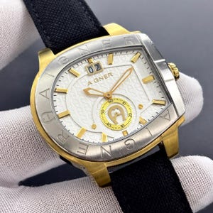 Puede incluir: Un reloj Aigner dorado y plateado con esfera blanca y correa negra. La esfera muestra la fecha "02" y el nombre de la marca "AIGNER". El reloj tiene agujas e indicadores horarios dorados.