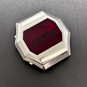 Può includere: Un oggetto geometrico color argento con un centro rettangolare rosso scuro. L'oggetto ha un design a cerniera e una finitura metallica lucida. La forma generale è un ottagono con bordi smussati.