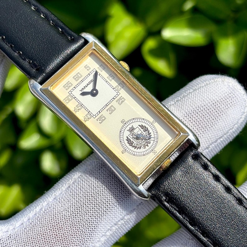 May include: Rectangular gold-toned wristwatch with a black leather band. The watch face is cream-colored with black numerals and hands. The watch face has the word "YANASE" and a crest design. The watch is set against a blurred green background.