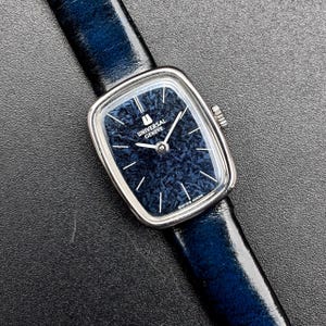 Op de afbeelding: Een Universal Geneve polshorloge met een rechthoekige zilverkleurige kast en een getextureerde donkerblauwe wijzerplaat. Het horloge heeft zilverkleurige wijzers en uurmarkeringen, een kroon aan de rechterkant en een glanzende donkerblauwe lederen band.