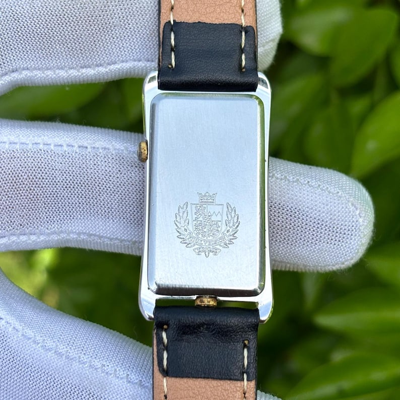 May include: Close-up of a rectangular silver watch case with a black leather strap. The watch back features a crest design. The watch band is black with a tan underside.