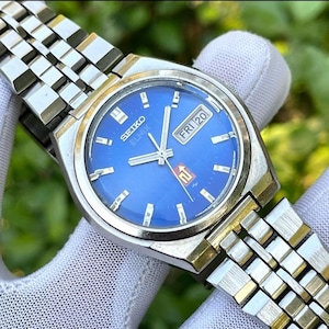 May include: A vintage Seiko Elnix wristwatch with a vibrant blue dial. The watch features a silver-tone case and a stainless steel band. The dial displays the day and date, with the day reading "FRI 20".