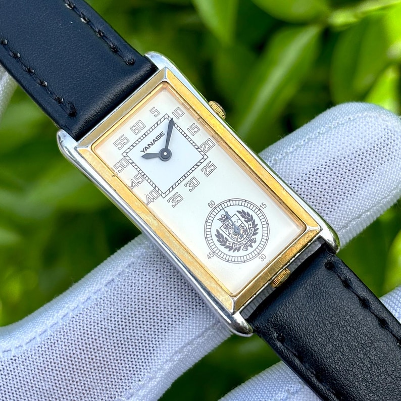 May include: Rectangular wristwatch with a gold-tone bezel and a white face. The watch features black hands, a black leather band, and the brand name "YANASE" is visible. The watch face also displays a crest and minute markers.