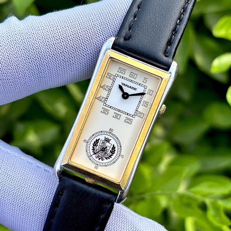 May include: A rectangular gold-toned wristwatch with a black leather band. The watch face is cream-colored with black numerals and hands. The brand name "YANASE" is displayed. A circular emblem is at the bottom of the watch face.