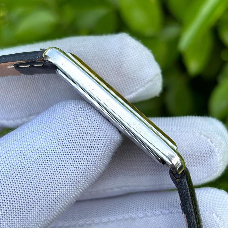 May include: A rectangular silver wristwatch with a black leather band. The watch has a minimalist design with a silver case and a black strap. The watch is held by a gloved hand.