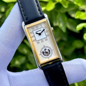 May include: A rectangular wristwatch with a gold-colored frame and a black leather band. The watch face is cream-colored with black numerals and hands. The brand name "YANASE" is visible. The watch is held by a gloved hand.