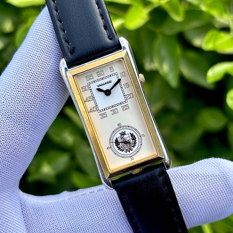 May include: A rectangular wristwatch with a gold-colored frame and a black leather band. The watch face is cream-colored with black numerals and hands. The brand name "YANASE" is visible. The watch is held by a gloved hand.