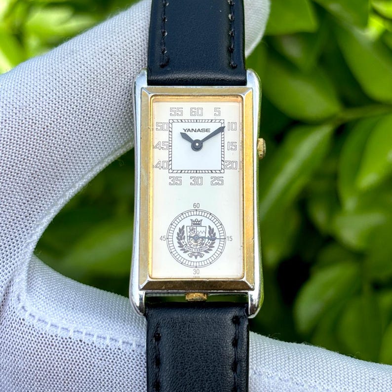May include: A rectangular wristwatch with a gold-tone bezel and a white face. The watch features a black leather band and the brand name "YANASE" is visible. The watch face displays numbers and a crest design.