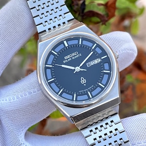 Puede incluir: Un reloj Seiko Solar Quartz plateado con una esfera azul oscuro y una correa plateada a juego. La esfera del reloj muestra el día y la fecha, con el logotipo de Seiko y las palabras "Solar Quartz" visibles. El reloj tiene una caja rectangular y una correa de eslabones.