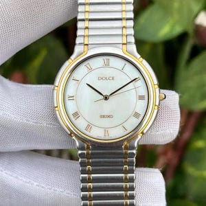 Puede incluir: Un reloj Seiko Dolce plateado y dorado con esfera blanca y agujas negras. El reloj presenta números romanos como marcadores de hora y el nombre de la marca se muestra en la esfera. La correa del reloj es plateada con detalles dorados.