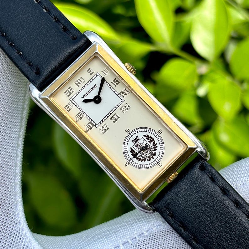 May include: A rectangular wristwatch with a white face, gold-colored bezel, and black leather band. The watch face displays the brand name "YANASE" and a circular emblem. The watch is set against a blurred green background.