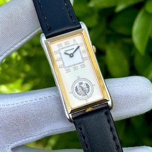 May include: Rectangular wristwatch with a gold-tone case and a black leather band. The watch face is white with black numerals and hands, and the brand name "YANASE" is visible. A decorative emblem is at the bottom of the watch face.