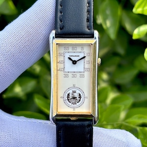 May include: A rectangular wristwatch with a gold-colored frame and a black leather band. The watch face is white with black numerals and the brand name "YANASE". The watch has a second dial at the bottom and a black leather band.