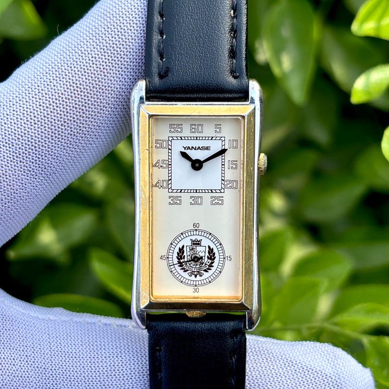 May include: A rectangular wristwatch with a gold-colored frame and a black leather band. The watch face is white with black numerals and the brand name "YANASE". The watch has a second dial at the bottom and a black leather band.
