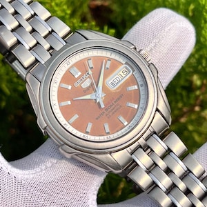 May include: A stainless steel Seiko 5 automatic wristwatch with a copper-colored dial. The watch features a silver band, Roman numeral hour markers, and a day-date display. The watch is water resistant.