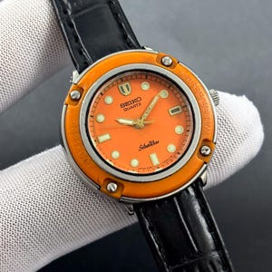 Może przedstawiać: Zegarek Seiko Quartz SilverWave w stylu vintage z pomarańczową tarczą i ramką. Zegarek ma złote wskazówki i znaczniki, czarny skórzany pasek, a widoczny jest napis "WATER RESIST". Klasyczny zegarek.