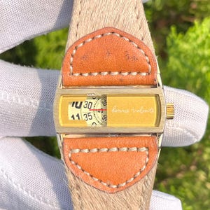 May include: A vintage wristwatch with a gold-toned rectangular face and a tan leather strap. The watch face displays the time and the words "bonne volonte". The strap is made of a light brown material with orange leather accents.