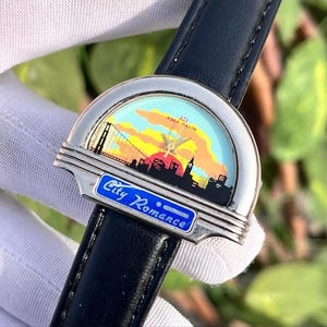 May include: A wristwatch with a black leather band and a silver-toned face. The watch face features a colorful cityscape design with a bridge and the text "City Romance". The watch hands point to the time.