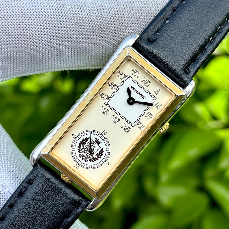 May include: A rectangular wristwatch with a gold-toned frame and a black leather band. The watch face is cream-colored with black numerals and hands. The brand name "YANASE" is visible on the watch face. The watch has a second dial with a crest.
