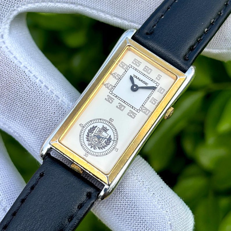 May include: A rectangular wristwatch with a gold-colored frame and a white face. The watch has a black leather band. The face displays the brand name "YANASE" and a circular emblem. The watch measures time with black hands and numerals.