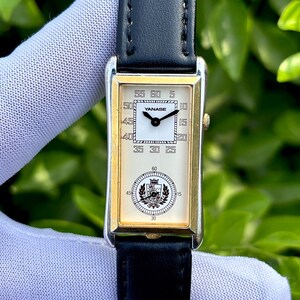 May include: Rectangular gold-toned wristwatch with a black leather band. The watch face is white with black numerals and hands. The brand name "YANASE" is visible. The watch has a classic, elegant design.