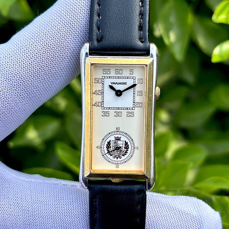 May include: Rectangular gold-toned wristwatch with a black leather band. The watch face is white with black numerals and hands. The brand name "YANASE" is visible. The watch has a classic, elegant design.
