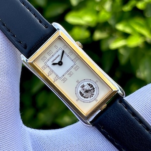 May include: A rectangular wristwatch with a gold-colored bezel and a black leather band. The watch face is white with black numerals and hands. The brand name "YANASE" is visible. The watch has a classic, elegant design.