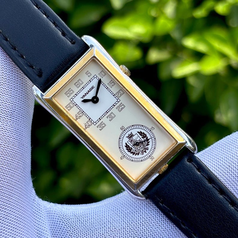 May include: A rectangular wristwatch with a gold-colored bezel and a black leather band. The watch face is white with black numerals and hands. The brand name "YANASE" is visible. The watch has a classic, elegant design.