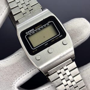 Puede incluir: Un reloj digital Casio Quartz plateado con una pantalla negra rectangular y una correa plateada a juego. La esfera del reloj muestra el logotipo de Casio y los días de la semana. El reloj tiene tres botones a la derecha y uno a la izquierda.