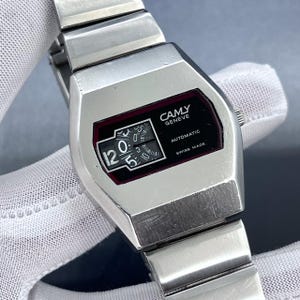Op de afbeelding: Een vintage zilverkleurig CAMY Geneve automatisch polshorloge. De wijzerplaat is zwart met witte cijfers en wijzers, en de tekst "CAMY GENEVE", "AUTOMATIC" en "SWISS MADE". De horlogeband is ook zilverkleurig.