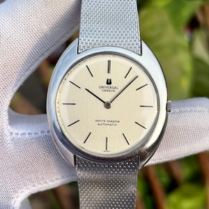 May include: A silver Universal Geneve White Shadow Automatic wristwatch with a cream face and a mesh strap. The watch face has black stick hour markers and the brand name. The watch is shown against a white background.
