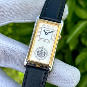 May include: Rectangular wristwatch with a gold-tone case and a white face. The watch features black hands, a black leather band, and the brand name "YANASE" on the face. The watch has a second dial with a crest.